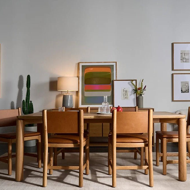 Modern dining room with wooden table and six tan leather chairs, neutral rug, cactus, bookshelf, framed art, lamp, and boucle accent chair with pillow, creating a warm and stylish space.