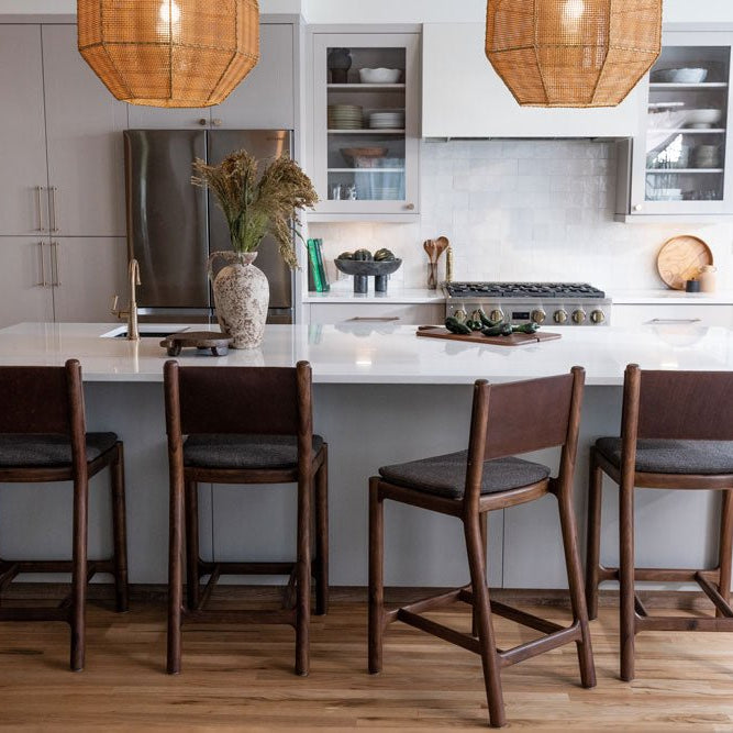 Modern kitchen with a white island and four Denver Modern Avon Counter Stools in dark wood frames with upholstered seats, under woven pendant lighting, surrounded by light wood flooring and sleek cabinetry.
