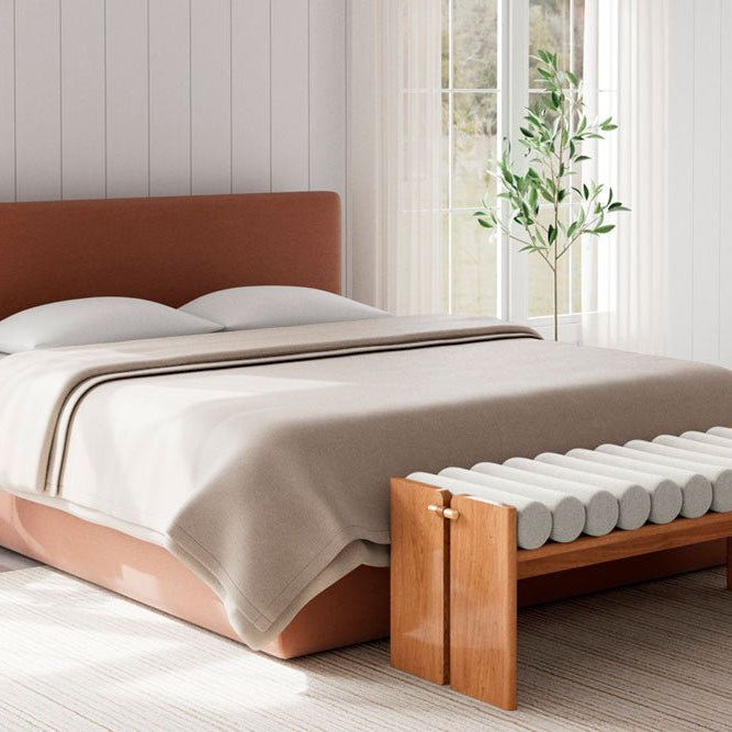 Modern bedroom with a terracotta-upholstered bed and a sculptural wood-and-fabric bench at the foot, set in a bright room with white paneled walls and natural light streaming in.