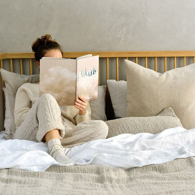 Woman sitting comfortably on a bed with light neutral bedding, reading a book titled “Words.” The bed features a natural wood spindle headboard, layered with large beige pillows and a cozy knit blanket.