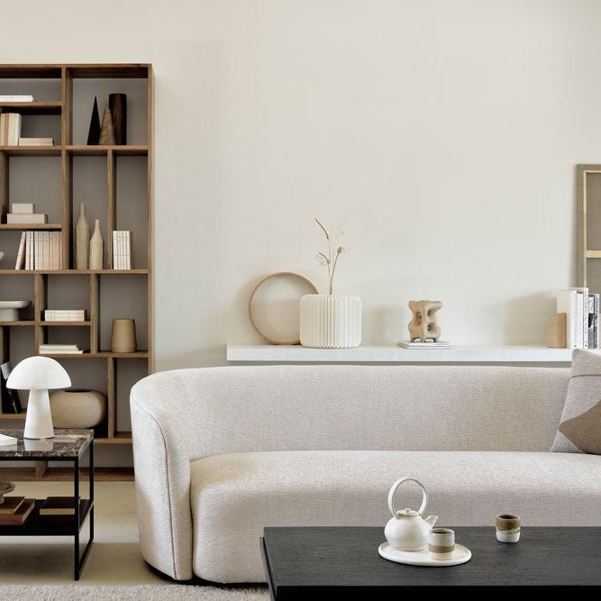 Modern living room with a tall open wooden bookcase styled with books and decorative objects, paired with a curved cream sofa, marble side table, and black coffee table.