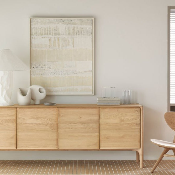 Light oak Denver Modern sideboard with four panel doors, styled with a sculptural white lamp, modern ceramic decor, and abstract artwork above, paired with a matching wood accent chair in a bright, minimalist room.