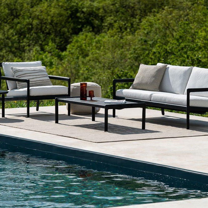 Modern black-framed outdoor sofa set with white cushions and accent pillows, placed beside a pool with scenic green hills in the background.
