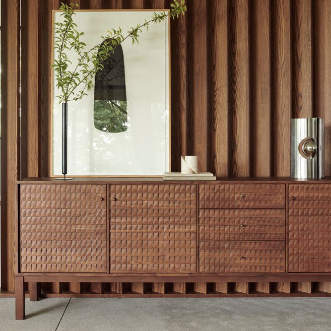 Modern wooden storage cabinet with a textured front, styled with minimalist decor including a framed abstract print, branch arrangement, and metallic vase, set against a vertical wood-paneled wall.