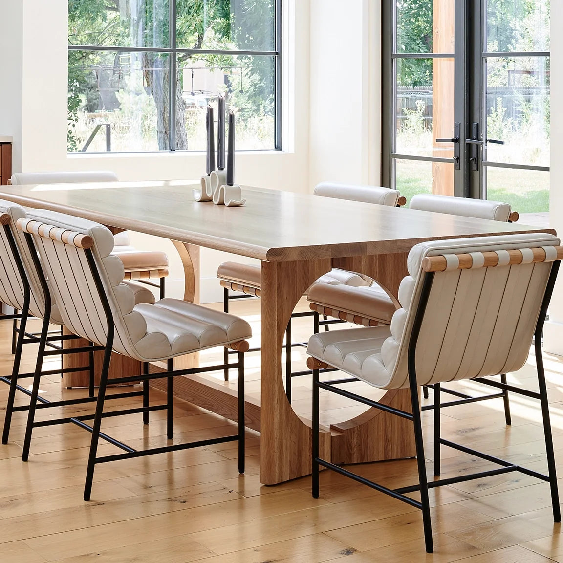 Modern dining room with a light wood table and upholstered dining chairs featuring strap detailing, set in a bright space with large windows.