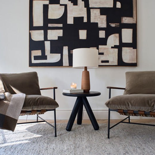 Two Denver Modern Vail Lounge Chairs with leather straps and plush olive cushions sit beside a black round side table with a wooden lamp, candle, and book, framed by abstract wall art in a bright, modern living room.