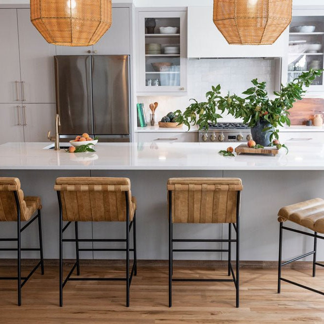 Modern kitchen with a white island and four woven leather bar stools on black metal frames, styled with natural wood floors and greenery, part of Denver Modern’s Vail Series collection.