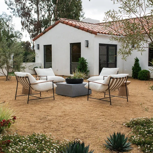An inviting outdoor seating area with stylish cream-colored chairs arranged around a central fire pit, set against a backdrop of a white stucco house with terra cotta roof tiles, surrounded by an array of blooming flowers and desert plants.