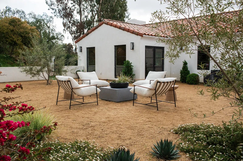 An inviting outdoor seating area with stylish cream-colored chairs arranged around a central fire pit, set against a backdrop of a white stucco house with terra cotta roof tiles, surrounded by an array of blooming flowers and desert plants.