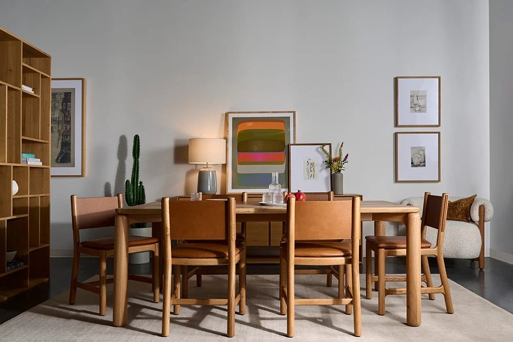 Modern dining room with wooden table and six tan leather chairs, neutral rug, cactus, bookshelf, framed art, lamp, and boucle accent chair with pillow, creating a warm and stylish space.