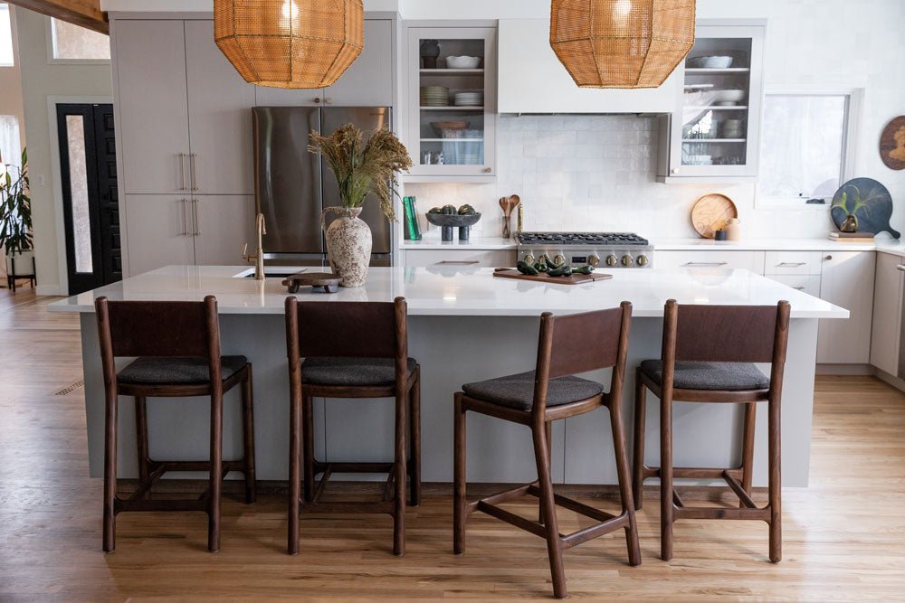 Modern kitchen with a white island and four Denver Modern Avon Counter Stools in dark wood frames with upholstered seats, under woven pendant lighting, surrounded by light wood flooring and sleek cabinetry.
