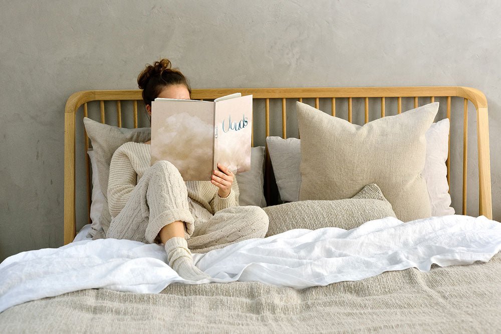 Woman sitting comfortably on a bed with light neutral bedding, reading a book titled “Words.” The bed features a natural wood spindle headboard, layered with large beige pillows and a cozy knit blanket.