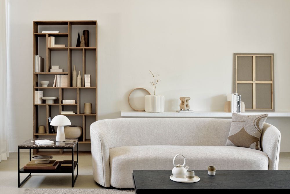 Modern living room with a tall open wooden bookcase styled with books and decorative objects, paired with a curved cream sofa, marble side table, and black coffee table.
