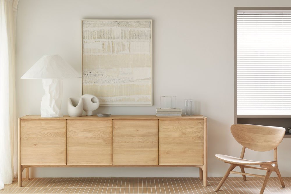 Light oak Denver Modern sideboard with four panel doors, styled with a sculptural white lamp, modern ceramic decor, and abstract artwork above, paired with a matching wood accent chair in a bright, minimalist room.