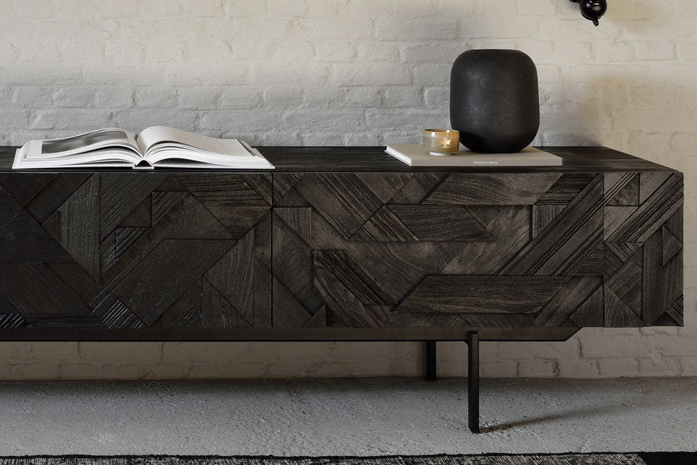 Dark wood Denver Modern media console with geometric inlay pattern, styled with an open book, black vase, and small cup, set against a white brick wall in a modern living space.
