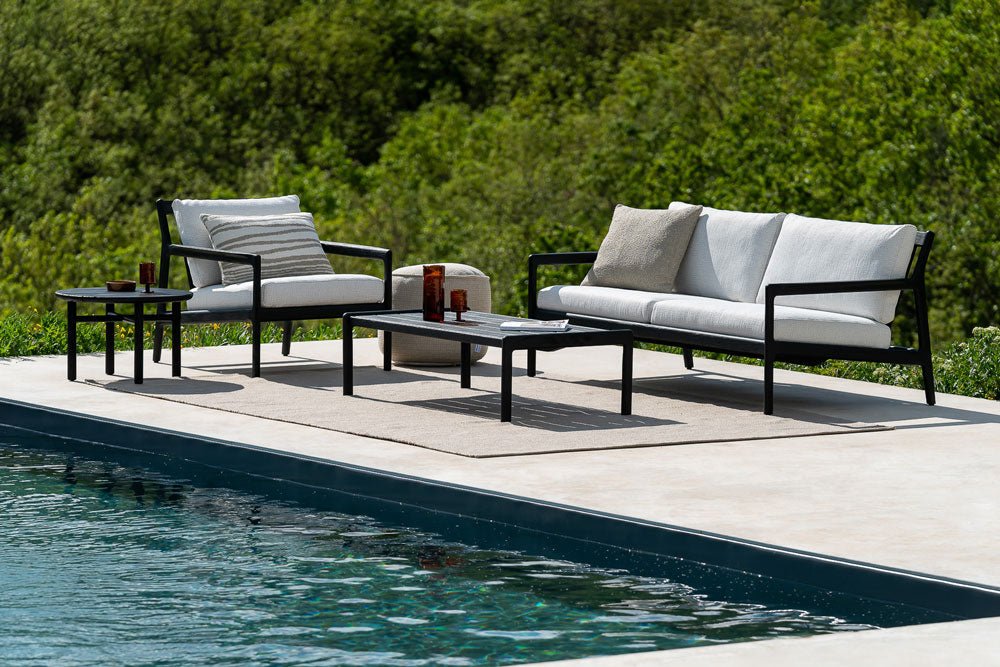 Modern black-framed outdoor sofa set with white cushions and accent pillows, placed beside a pool with scenic green hills in the background.