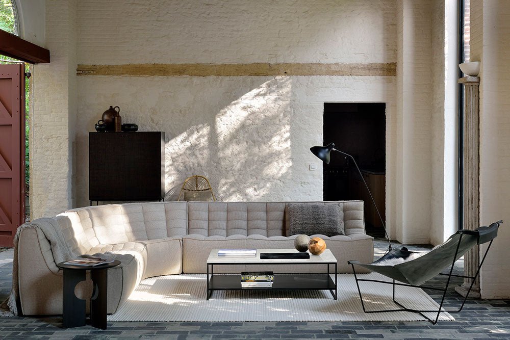 Spacious living room with a cream upholstered N701 modular sectional sofa, black metal coffee table, modern side table, and butterfly chair, set against textured white walls with tall windows.