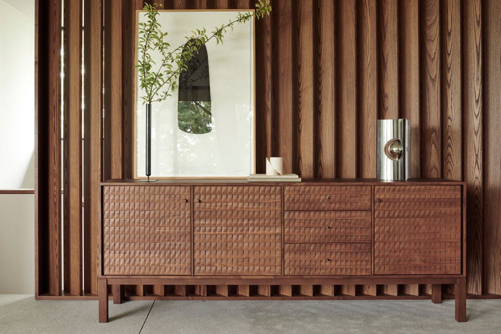 Modern wooden storage cabinet with a textured front, styled with minimalist decor including a framed abstract print, branch arrangement, and metallic vase, set against a vertical wood-paneled wall.