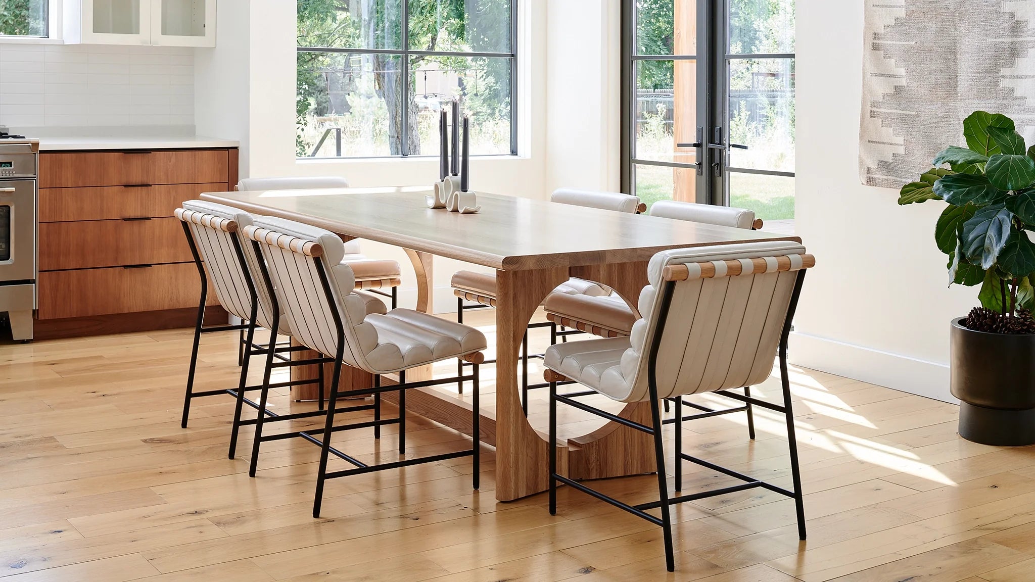 Modern dining room with a light wood table and upholstered dining chairs featuring strap detailing, set in a bright space with large windows.