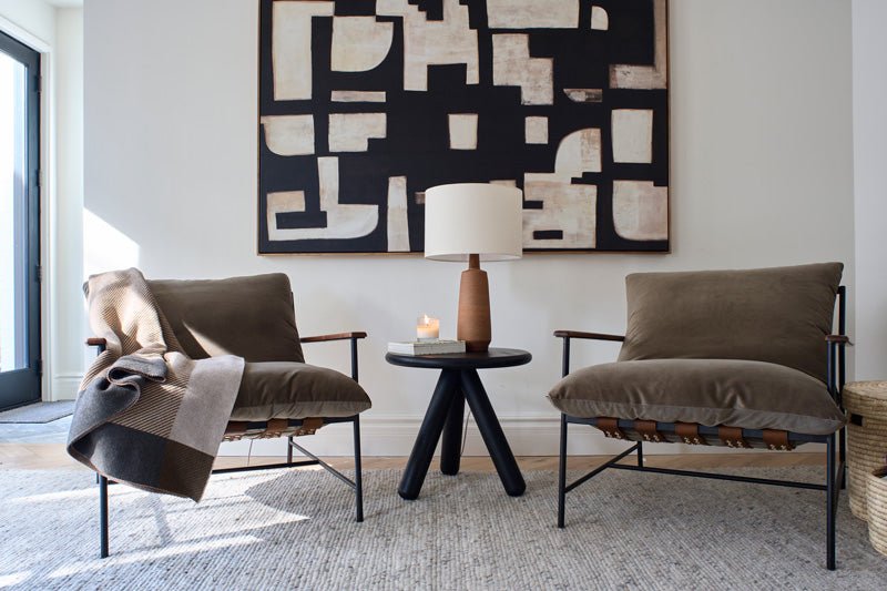 Two Denver Modern Vail Lounge Chairs with leather straps and plush olive cushions sit beside a black round side table with a wooden lamp, candle, and book, framed by abstract wall art in a bright, modern living room.