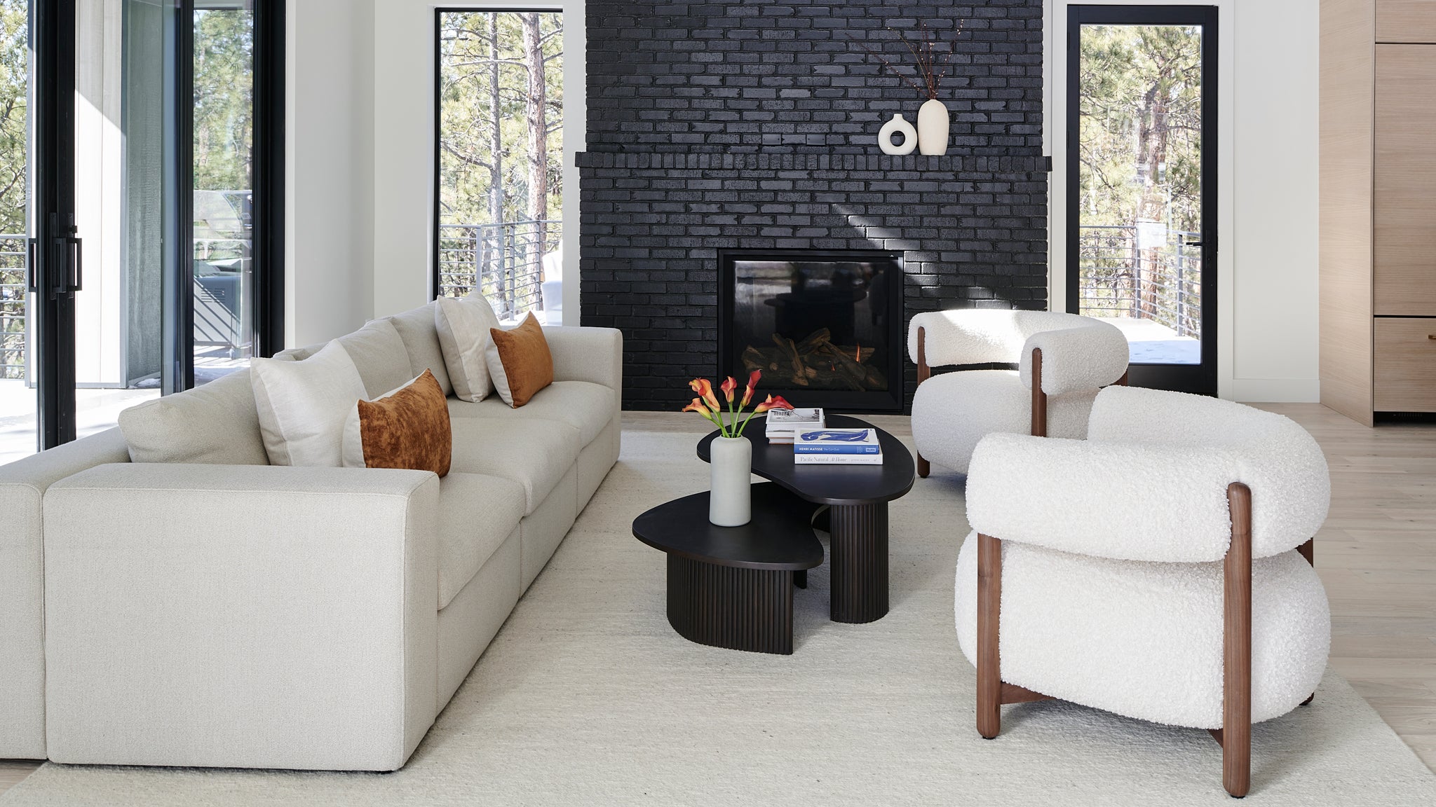 A stylish living room with a textured black fireplace, modern furniture, and forest views through large windows.