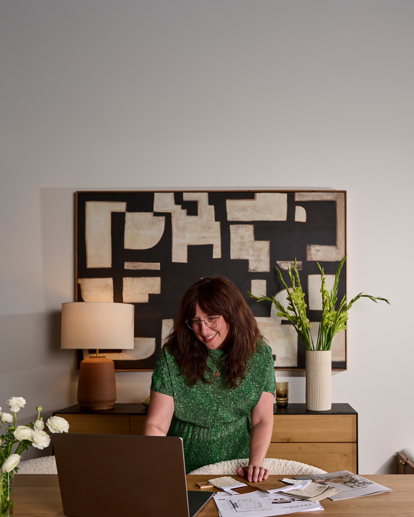 Woman sitting at a desk with a laptop, surrounded by decor elements like a lamp, plant, and abstract art.