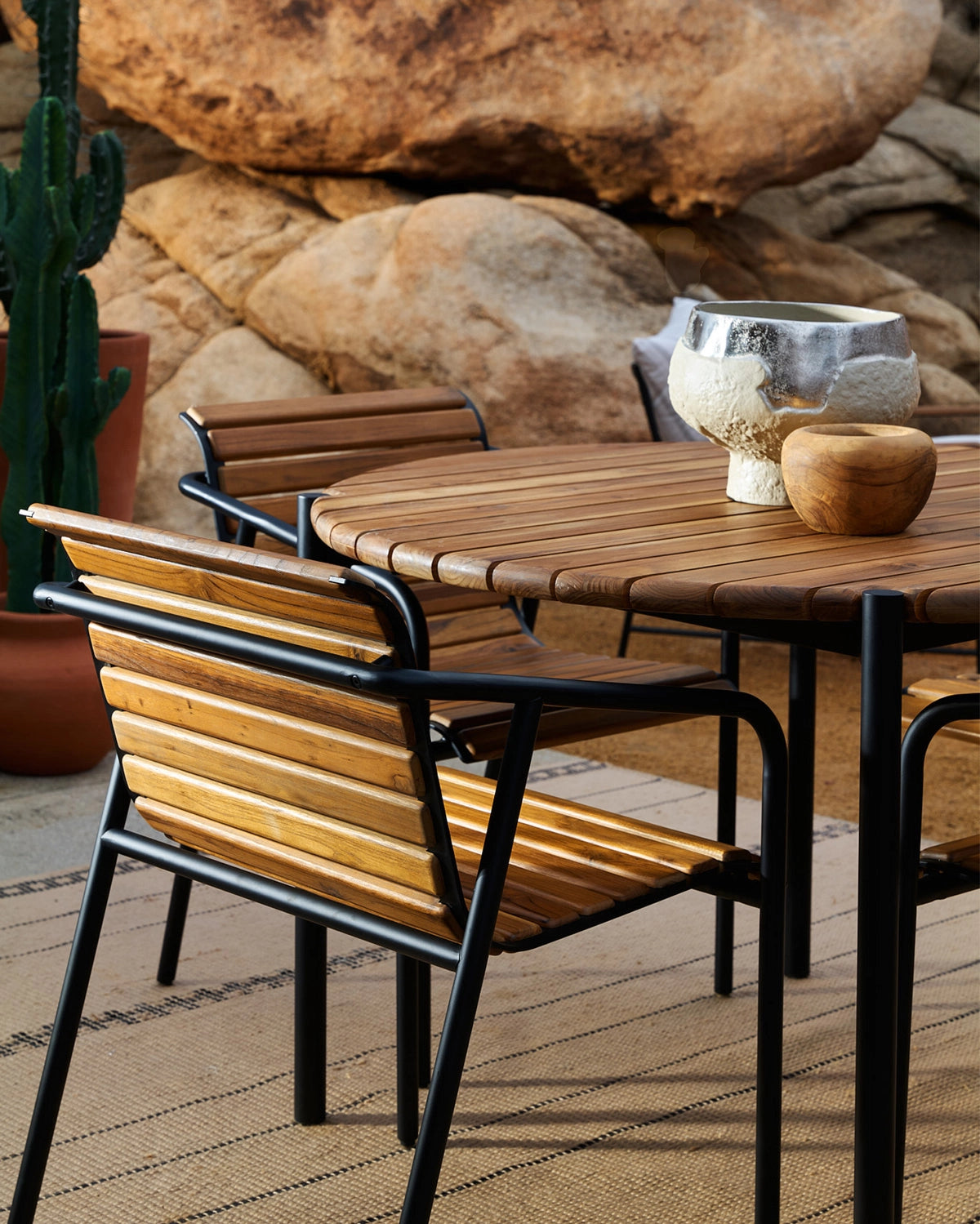 Outdoor Strata Round Dining Table styled with matching Strata dining chairs, featuring a round slatted wood top and black steel frame in a desert-inspired outdoor setting.