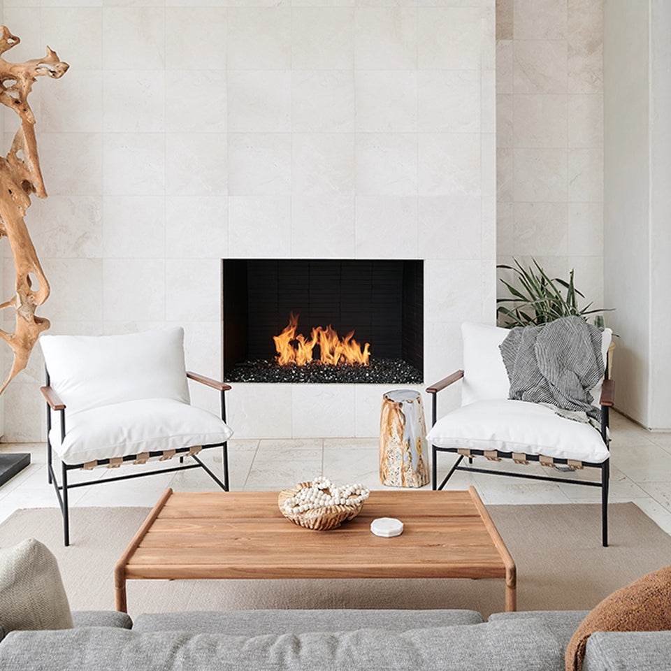 A spacious modern living area featuring a neutral-toned sofa with white cushions, a wooden coffee table, and a patterned rug. The space includes black-framed windows and doors, light wood flooring, and a mix of greenery and natural elements, creating a bright and inviting atmosphere.