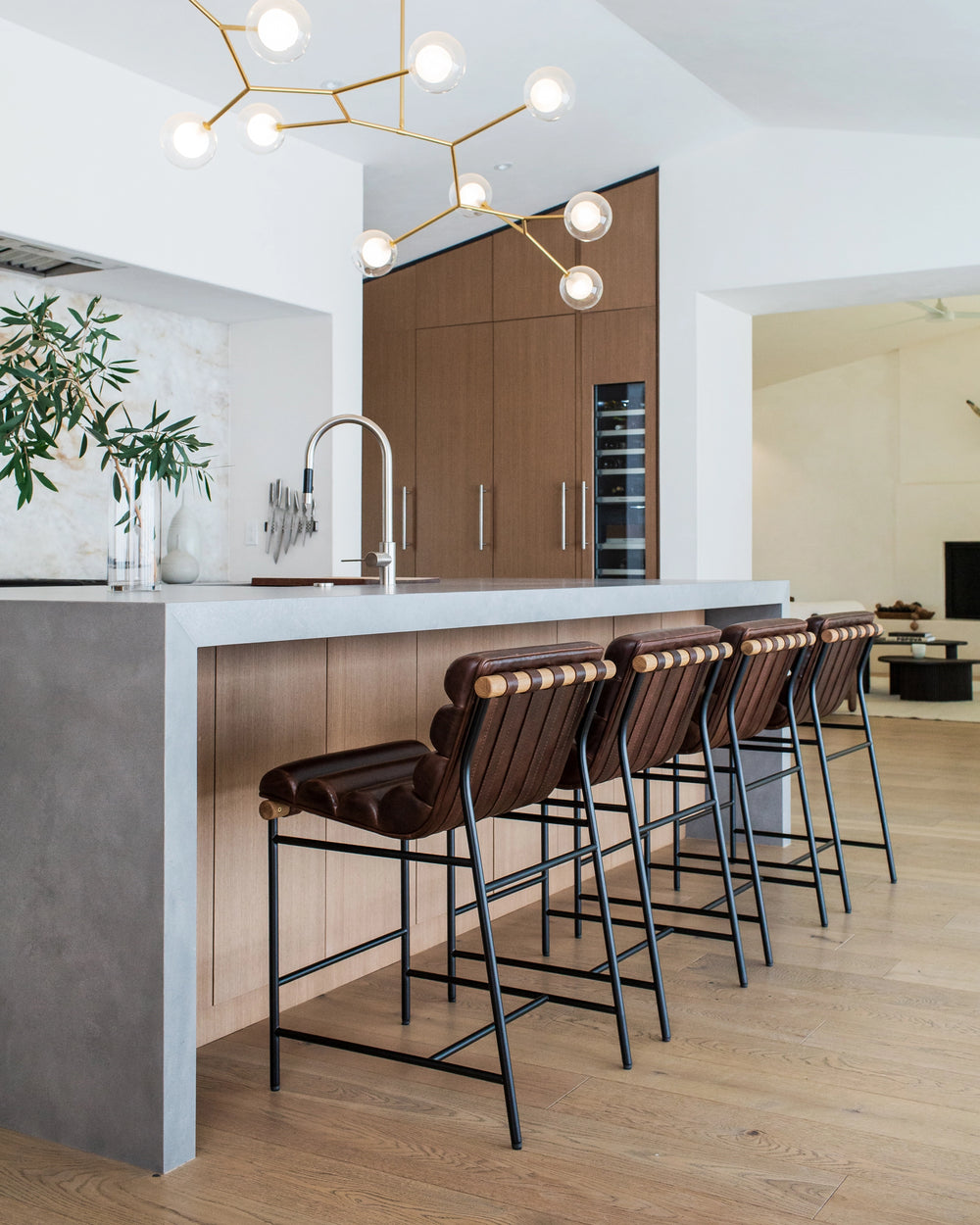 A contemporary kitchen setting with a sleek kitchen island, topped with a grey stone countertop and surrounded by a set of four Vail Stools with dark brown leather seats and black metal frames. 