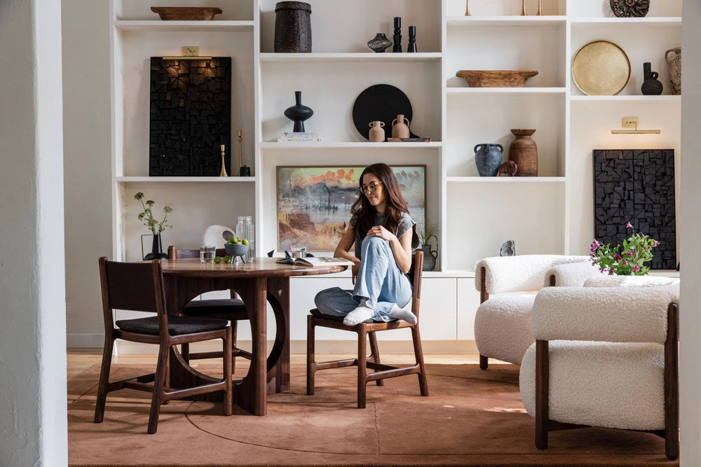 Woman sitting at a wooden dining table in a modern living room with white shelves and decor.
