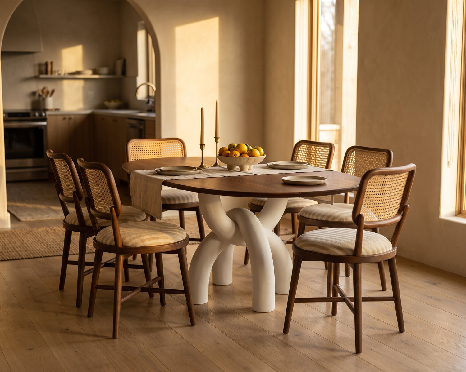 A dining set in a warm, sunlit room featuring a round wooden table with a sculptural white base and six matching wooden chairs with woven cane backs and cream cushions.