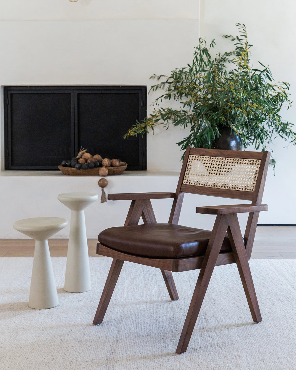 A contemporary room featuring a dark wooden Lark Accent Chair with a woven cane backrest and a smooth brown leather seat. The chair is accompanied by two cream-colored, cone-shaped side stools, adding a modern touch to the space.