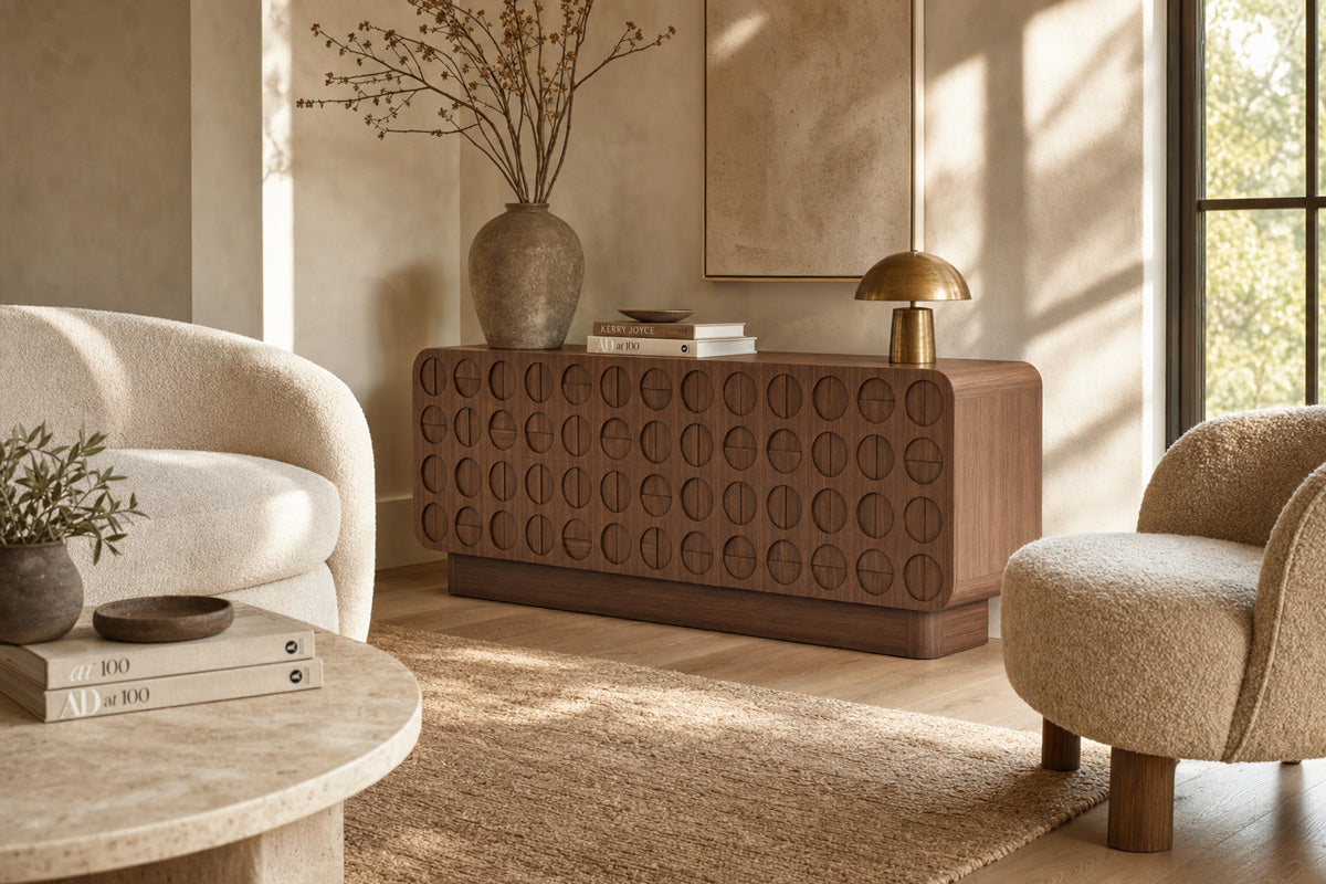 A mid-century modern wooden sideboard or credenza with a distinctive circular embossed pattern on the front, styled in a warm, sunlit living room with two textured armchairs and a marble coffee table.
