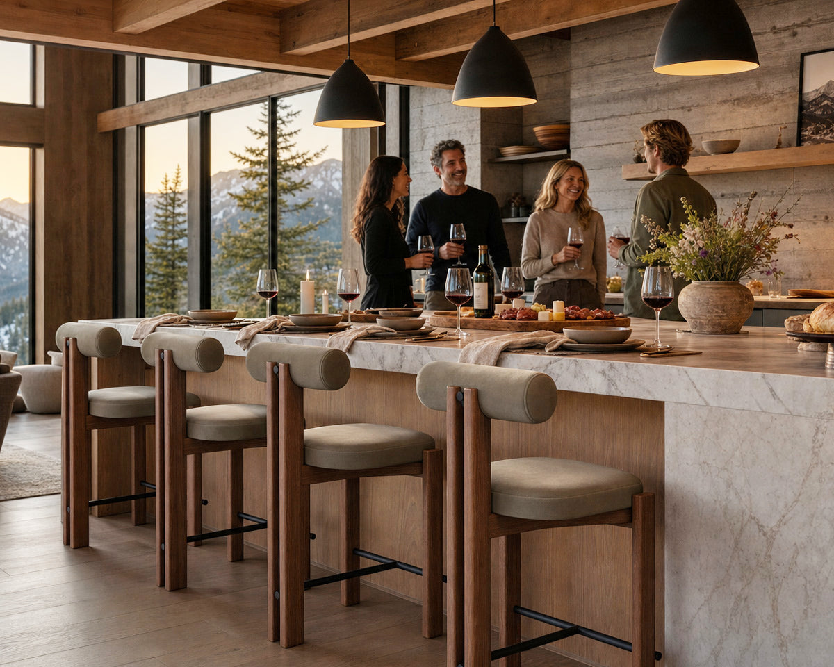 A modern kitchen island with a marble countertop and four wooden bar stools with beige cushions, set in a rustic mountain-view dining space with people standing nearby.