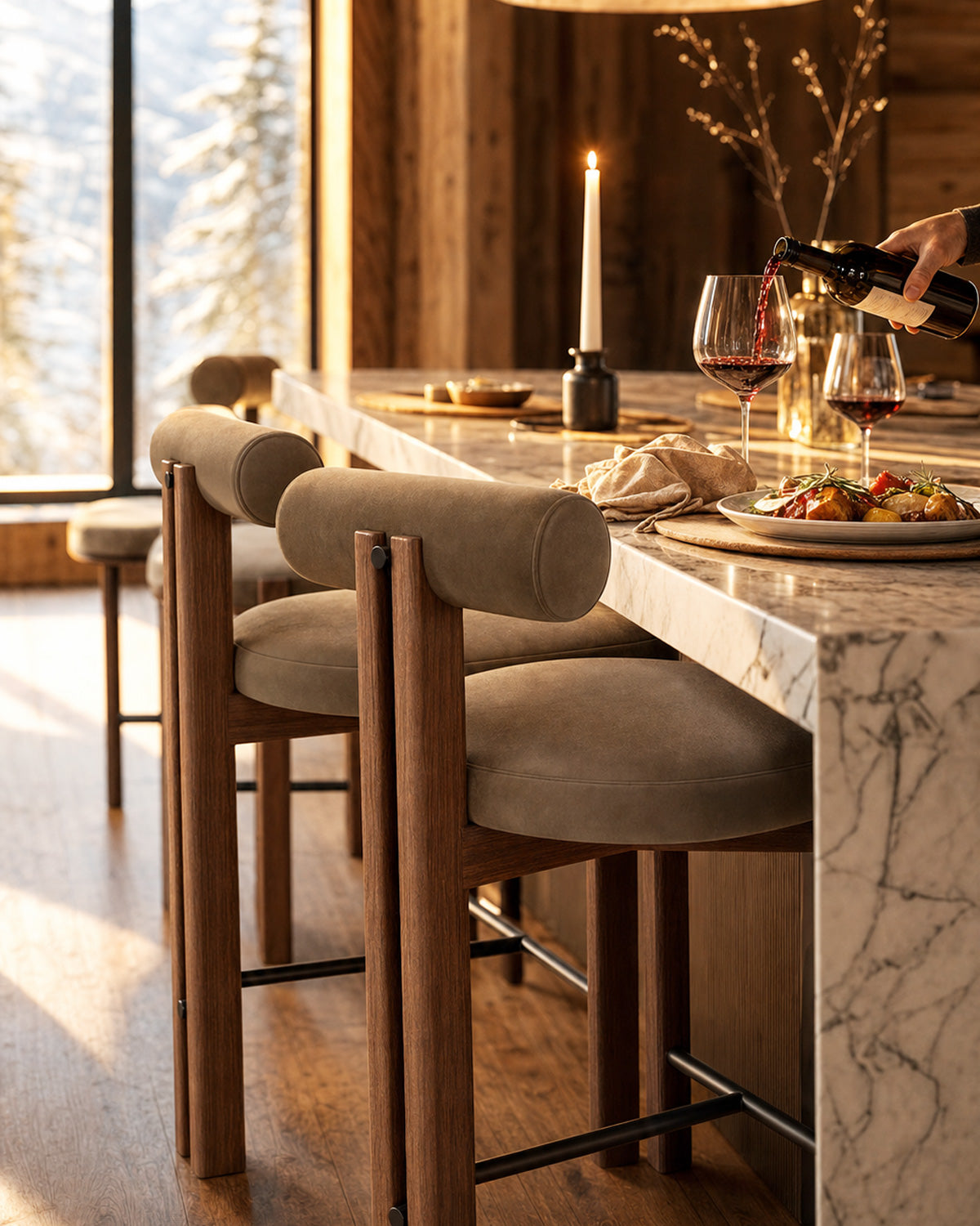A lifestyle scene in a warm, modern mountain chalet dining area featuring a set of bar stools with light beige velvet cushions and natural wood frames, set along a marble-topped kitchen island. A person is pouring red wine from a bottle into two wine glasses on the counter.