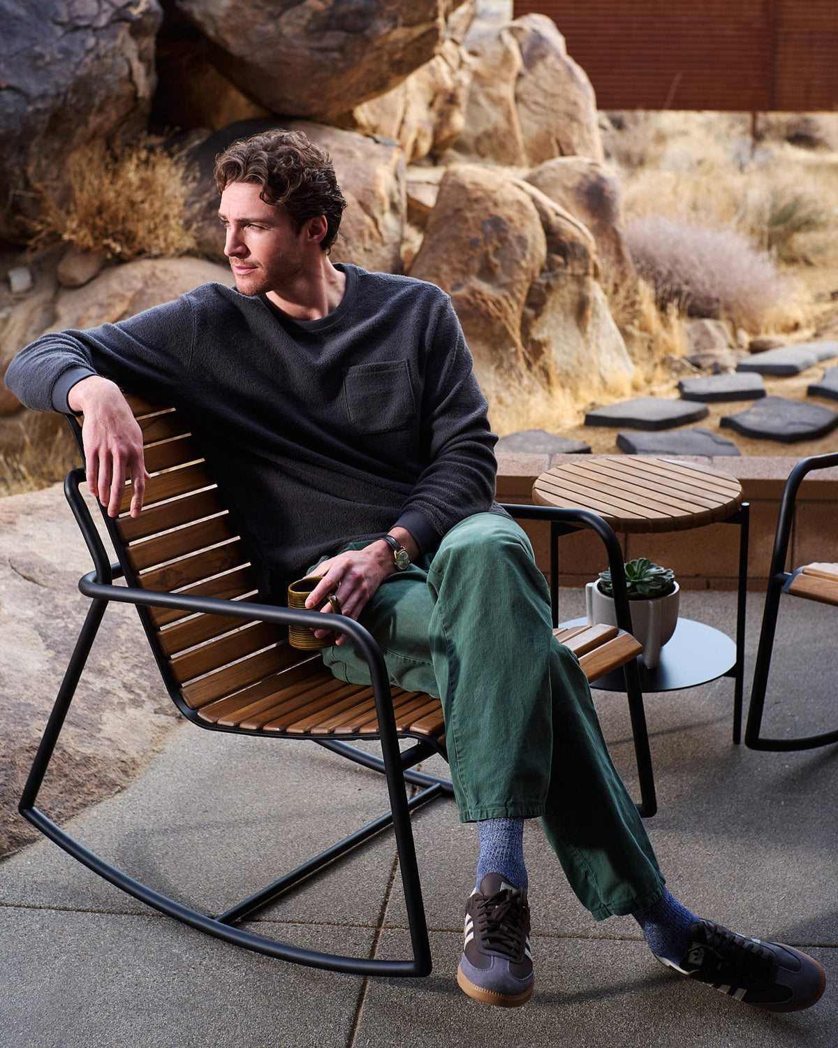 Outdoor Strata Rocker styled on a patio with a seated person, featuring a slatted wood seat, steel rocker base, and modern outdoor side tables