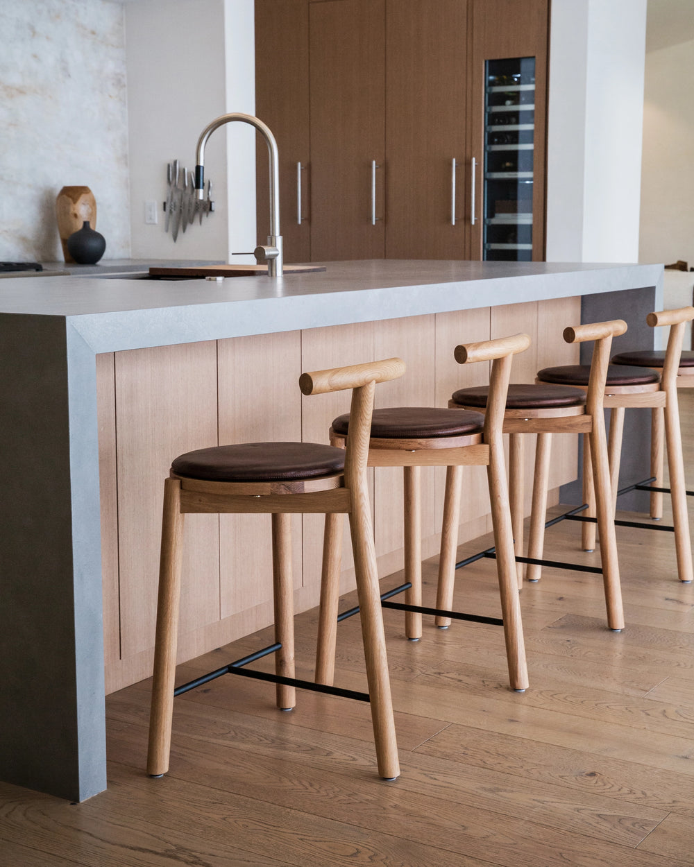 A minimalist kitchen interior featuring three Range Counter Stools in front of a kitchen island. The stools have a natural maple wood finish with slim legs, black footrests, and round leather cushions. 