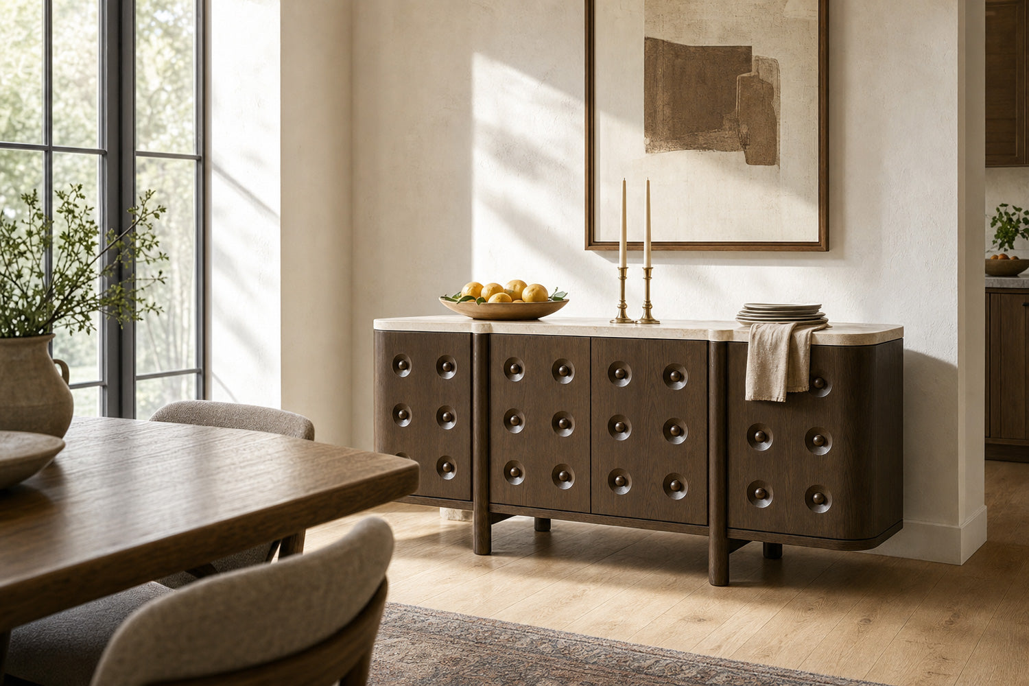 A modern sideboard or credenza in a warm brown finish with circular metal knobs, styled in a dining room setting with a wooden table, chairs, candles, and a bowl of lemons on top.