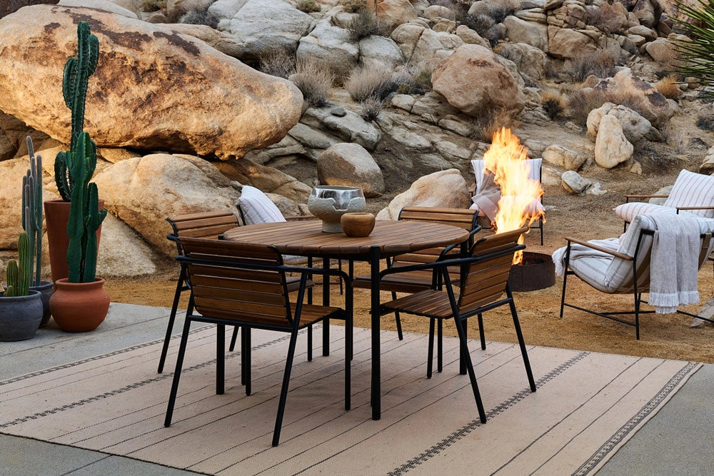 Outdoor patio setting with a Strata Round Dining Table and Strata Dining Chairs around a fire pit, surrounded by rocks and cacti.