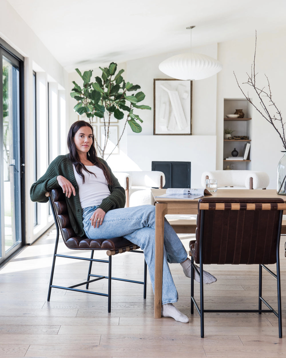 A bright dining room scene with a relaxed woman sitting in a brown leather Vail Dining Chair. She is casually dressed in a white top, green cardigan, and blue jeans, with her legs crossed. The dining table is made of light wood with contrasting dark stripes, matching the chair. 